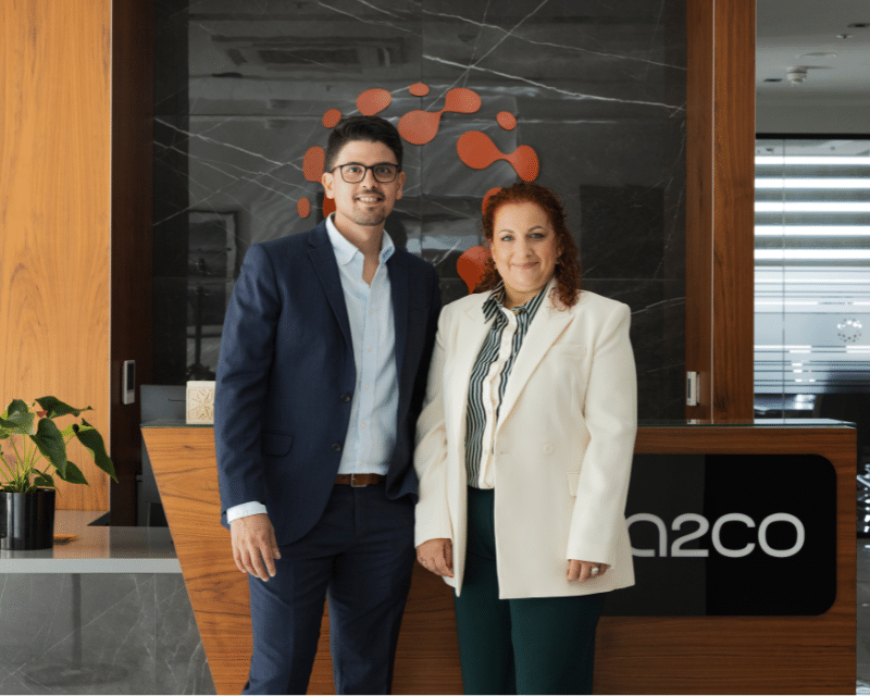 A2CO Partner's Clinton Cutajar and Antionette Scerri looking at the camera in front of an A2CO logo.