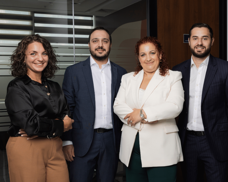 A2CO's Tax team including Partner Antoinette Scerri, Benjamin Zammit McKeon, Stephanie Bianco and Clivert Vella posing for a photo in front of a glass wall.