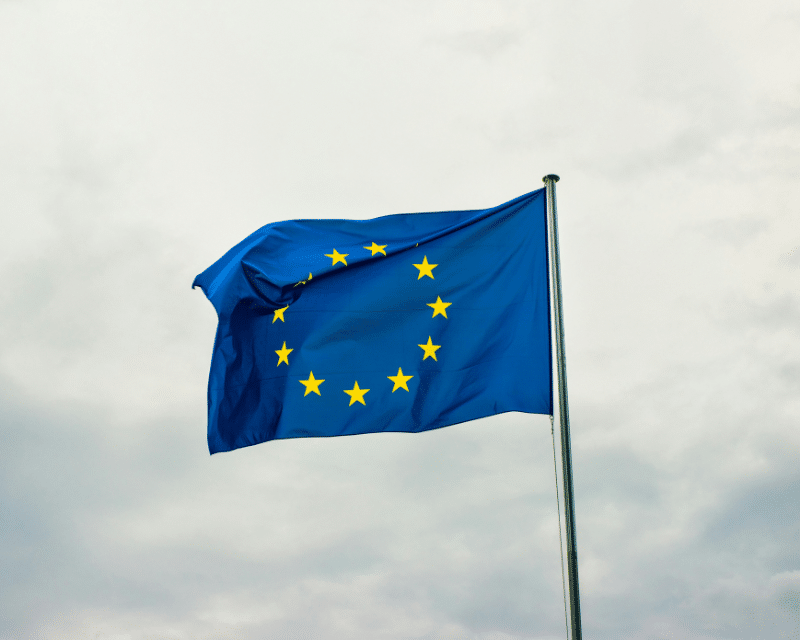 European Union flag with yellow stars waving on a flagpole against a cloudy sky.