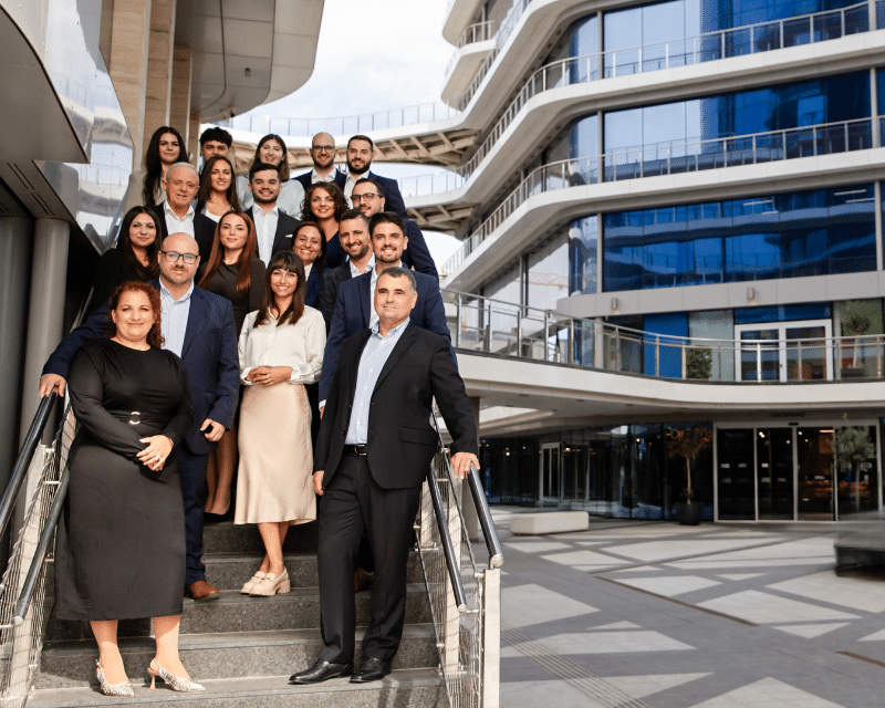 The A2CO team gathered on the outdoor staircase of their office building in Malta, posing together in professional attire.