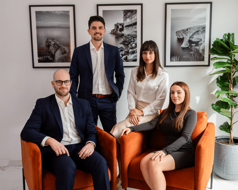A2CO team posing in a modern office lounge area with orange chairs and framed wall art. Clinton