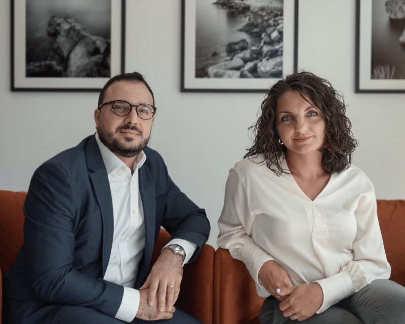 A2CO Senior Tax Managers, Stephanie Bianco and Benjamin Zammit McKeon, seated in an office setting with framed photographs on the wall behind them.