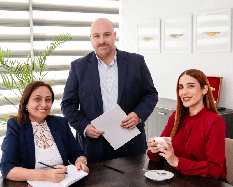 Three A2CO team members in a business meeting, smiling and engaged, with documents and coffee at the table.